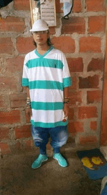 a young man wearing a striped shirt and a hat is standing in front of a brick wall .