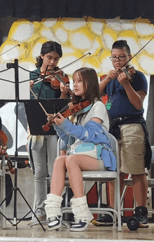 a girl sits in a chair playing a violin while two other girls play violins