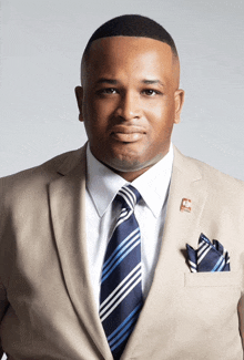 a man wearing a tan suit and a blue and white striped tie has a pocket square with the letter p on it