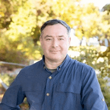 a man in a blue shirt is standing in front of a tree and smiling for the camera .