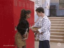 a man and a woman are standing next to each other in front of red lockers in a school hallway .