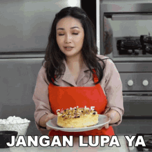 a woman in a red apron is holding a plate with a cake on it and the words jangan lupa ya above her