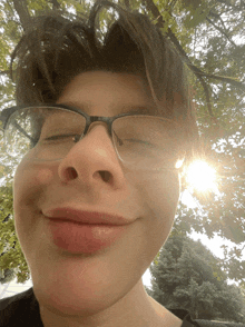 a young man wearing glasses is smiling with the sun shining through the trees in the background