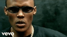 a man wearing sunglasses and a gold chain is standing in front of a black wall .