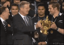 a man in a suit shakes hands with a man holding a trophy that says ' rugby ' on it