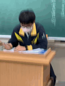 a man wearing a mask and glasses sits at a desk in front of a chalkboard with chinese writing on it