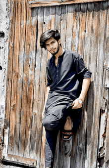 a young man leans against a wooden wall wearing a black shirt