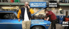 a man is talking on a cell phone while another man works on a car in a garage .