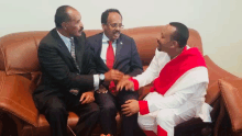 three men are sitting on a couch talking and one of them is wearing a red and white outfit