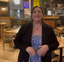 a woman wearing a black jacket and polka dot dress is standing in a restaurant with tables and chairs and smiling