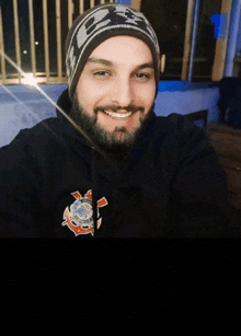 a man with a beard wearing a black hoodie and a beanie smiles for the camera