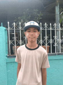 a young boy wearing a baseball cap with the letter h on it stands in front of a blue fence