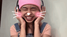 a woman wearing headphones and a pink hat has a cat face drawn on her cheeks