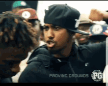 a man in a black hat is talking to another man in a crowd with proving grounds pg written on the bottom right