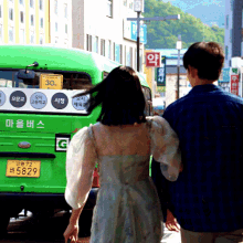 a man and woman are walking down a street in front of a green bus that says 30 on the back