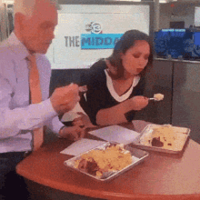 a man and a woman are sitting at a table eating food in front of a tv that says the midday