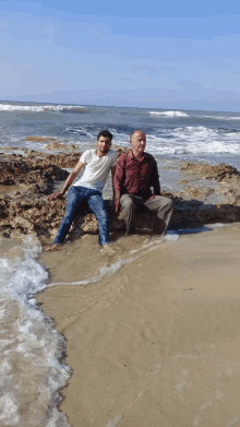 two men are sitting on a rock on the beach