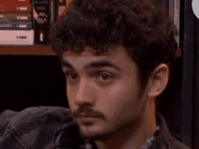 a man with curly hair and a beard is sitting in front of a bookshelf with books on it .