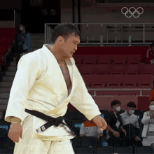 a man in a white kimono with a black belt is standing in a stadium with the olympic rings on the wall behind him