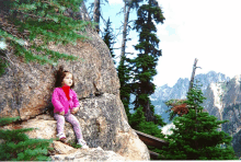 a little girl in a pink jacket sits on a rock in the woods