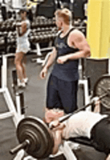 a man is lifting a barbell on a bench in a gym while a woman stands in the background .