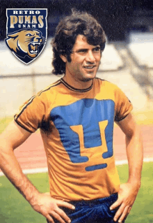 a man in a yellow pumas shirt stands on a field with his hands on his hips