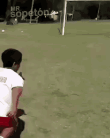 a man in a white shirt and red shorts is kneeling on a soccer field in front of a goal .