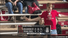 a woman in a red shirt stands in front of a scoreboard for a college football game between oklahoma st and texas tech