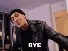 a man wearing glasses and a leather jacket is standing in a living room and saying bye .