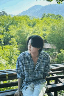 a man in a plaid shirt sits on a wooden railing with trees in the background