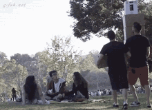 a group of people are sitting on the grass in a park while a man carrying a cardboard box stands in the background