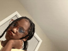 a young girl wearing glasses and braids looks at the camera with her hand on her chin