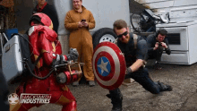 a man in an iron man suit holds a captain america shield in front of a sign that says hacksmith industries