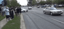 a group of people are standing on the side of the road watching a silver car drive down the street