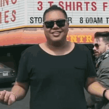 a man wearing sunglasses and a black shirt is standing in front of a sign that says t shirts .