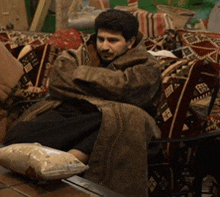 a man in a coat sits on a couch with a bag of rice on the floor