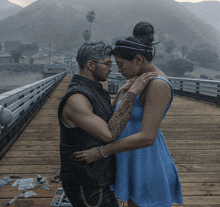 a man in a leather vest and a woman in a blue dress are hugging on a wooden bridge