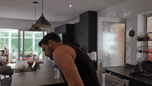 a man in a black tank top is squatting in a kitchen with a bottle of water on the counter