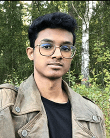 a man wearing glasses and a leather jacket stands in front of trees