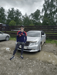 a young man stands in front of a silver car with a license plate that says ah 70