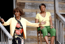 a woman in a yellow cardigan is standing next to a woman sitting in a chair