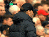 a man wearing a hat and a jacket stands in front of a crowd watching a soccer game between liv and tot