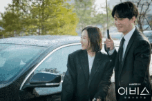 a man holding an umbrella stands next to a woman standing next to a car