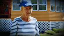 an elderly woman wearing a blue hat and striped shirt is standing in front of a house .