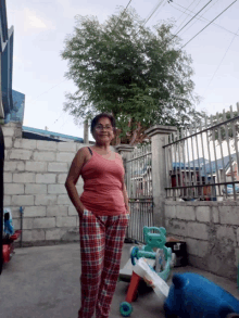 a woman wearing plaid pants and a pink tank top smiles in front of a fence