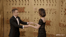 a man in a tuxedo and a woman in a black dress hold hands in front of a wall that says emmys