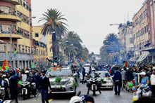 a group of people are walking down a street with a car decorated with green leaves