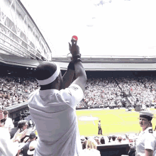 a man in a white shirt holds up a red ball in front of a crowd at a tennis stadium