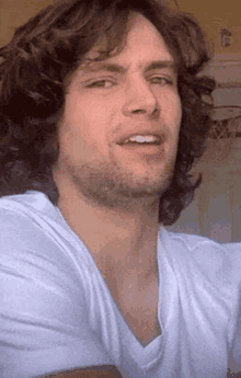 a man with long curly hair and a beard is wearing a white t-shirt and making a funny face .