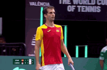 a man in a red and yellow shirt is holding a tennis racket in front of a sign that says the world cup of tennis